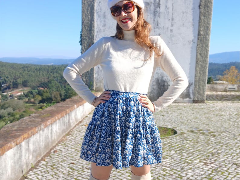 May include: A blue and white floral print skirt. The skirt is worn with a cream-coloured long-sleeved top. The outfit is completed with a white beret and sunglasses. The background features a stone wall and a scenic view.