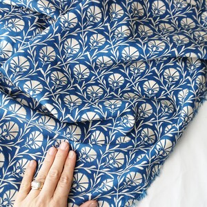 May include: A close-up of blue fabric with a white floral pattern. The design features stylized flowers and leaves, creating a repeating motif. The fabric has a soft, flowing appearance, suitable for various sewing projects.