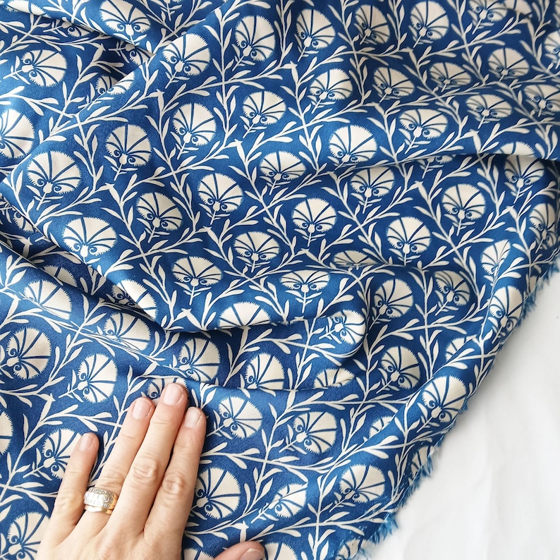 May include: A close-up of blue fabric with a white floral pattern. The design features stylized flowers and leaves, creating a repeating motif. The fabric has a soft, flowing appearance, suitable for various sewing projects.