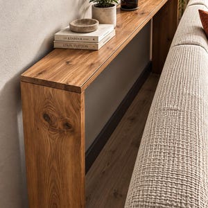 Puede incluir: Una mesa consola larga y estrecha hecha de madera natural con un patrón visible de vetas y nudos. La mesa cuenta con una superficie rectangular plana y dos paneles laterales verticales, que sostienen libros, un cuenco de piedra y una vela.