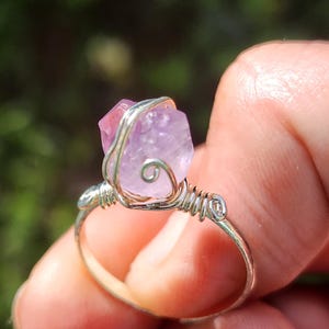 Op de afbeelding: Een handgemaakte ring met een ruwe, lichtpaarse amethist edelsteen. De steen wordt vastgehouden met zilverkleurig draad, wat een uniek en natuurlijk ontwerp creëert. De ringband is een eenvoudige, gepolijste zilverkleur, die de schoonheid van de edelsteen benadrukt.