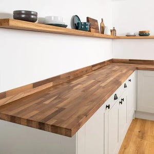 May include: A kitchen featuring a butcher block worktop made of light and dark brown wood strips, white cabinets with black round knobs, and a matching wooden floating shelf holding various ceramic bowls, plates, and wooden boards.