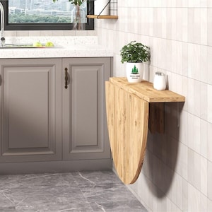 Op de afbeelding: Een aan de muur gemonteerde klaptafel van lichtgekleurd hout met een halfrond inklapbaar paneel. De tafel is bevestigd aan een betegelde muur en bevat een witte potplant met een groene boomafbeelding en een kleine witte cilindrische houder.