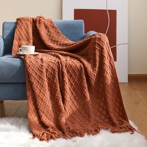 Könnte beinhalten: Eine braune gestrickte Wolldecke mit Rautenmuster und Fransen. Die Decke liegt auf einem blauen Sofa.
