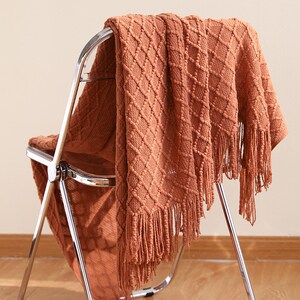 Könnte beinhalten: Eine braune gestrickte Wolldecke mit Rautenmuster und Fransen. Die Decke liegt über einem Metallstuhl.