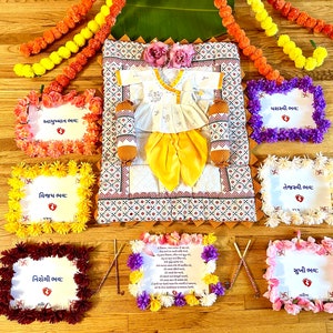 May include: A white baby outfit with a yellow dhoti and a white embroidered top, laid out on a white and brown patterned fabric. The outfit is surrounded by small framed signs with floral decorations and Gujarati text. The signs read "Yashvi Bhav", "Ayushman Bhav", "Vijay Bhav", "Nirogi Bhav", "Tejasvi Bhav", and "Sukhi Bhav".