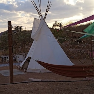 May include: A white teepee tent with a brown hammock hanging in front of it. The teepee is set up in a desert landscape with rocks and trees in the background.