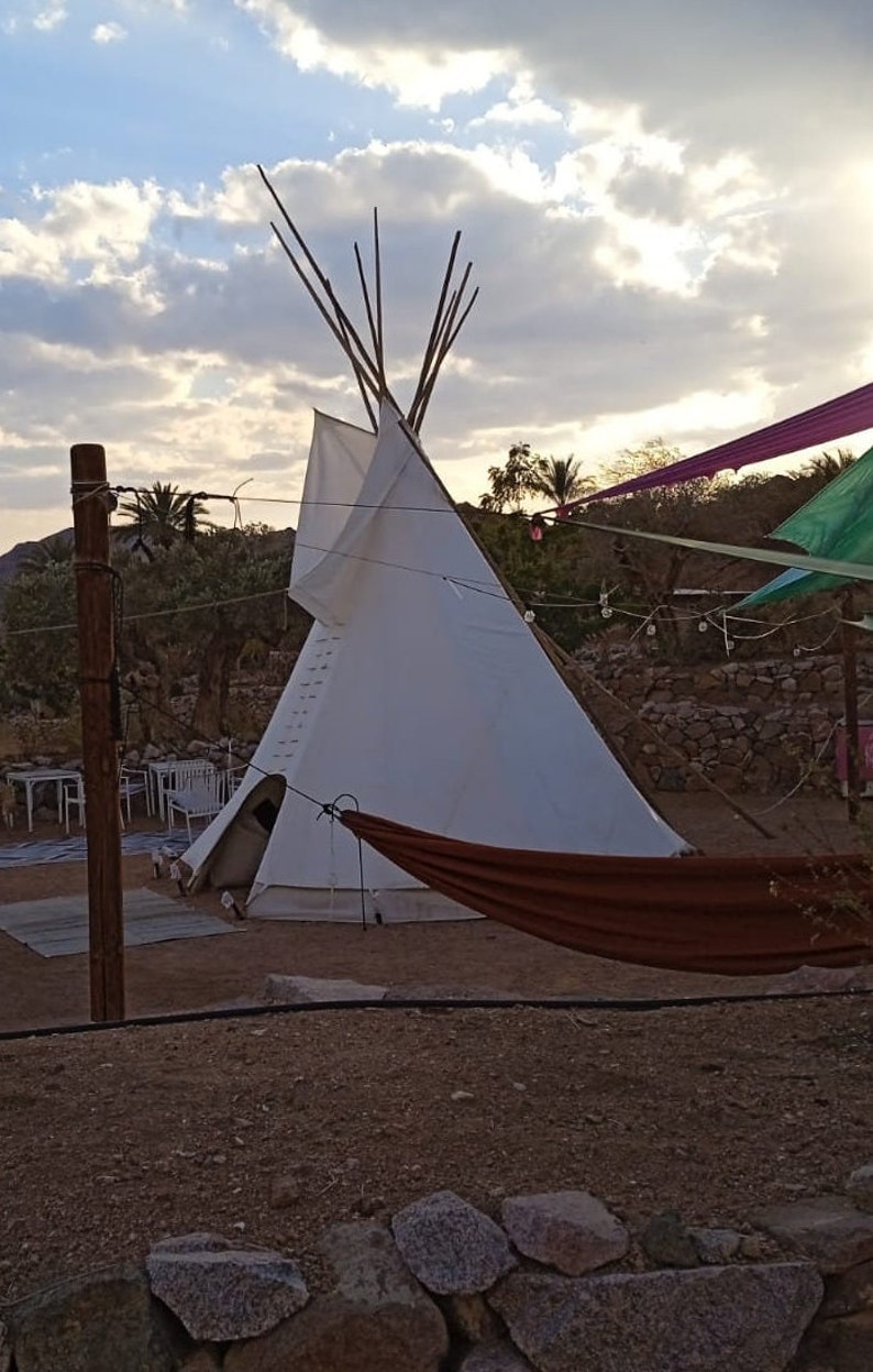 May include: A white teepee tent with a brown hammock hanging in front of it. The teepee is set up in a desert landscape with rocks and trees in the background.