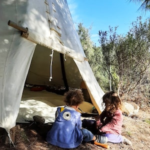 May include: Two children playing inside a white teepee tent. The girl is wearing a pink shirt and the boy is wearing a blue and yellow hooded sweatshirt.