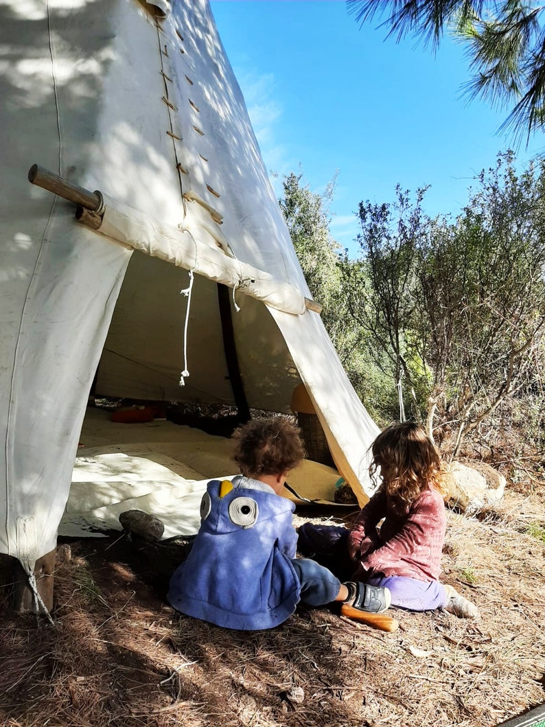 May include: Two children playing inside a white teepee tent. The girl is wearing a pink shirt and the boy is wearing a blue and yellow hooded sweatshirt.