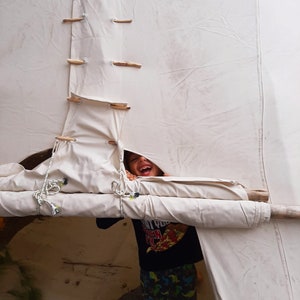 May include: A child peeks out from behind a white canvas tent, smiling and wearing a black shirt with a pizza graphic and green and black patterned pants.