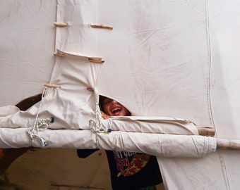 May include: A child peeks out from behind a white canvas tent, smiling and wearing a black shirt with a pizza graphic and green and black patterned pants.