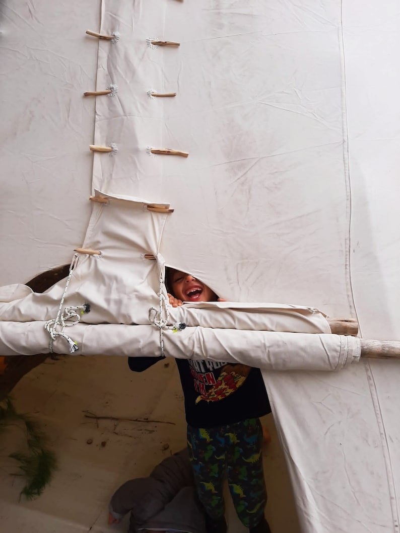 May include: A child peeks out from behind a white canvas tent, smiling and wearing a black shirt with a pizza graphic and green and black patterned pants.