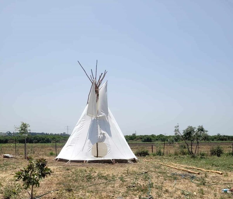 May include: A white teepee tent with a wooden frame, set up in a grassy field. The tent has a flap open at the front, revealing a dark interior.