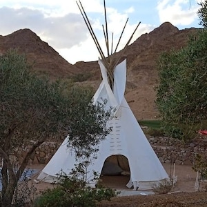 May include: A white teepee tent with a wooden frame, set up outdoors against a backdrop of brown hills and green trees.