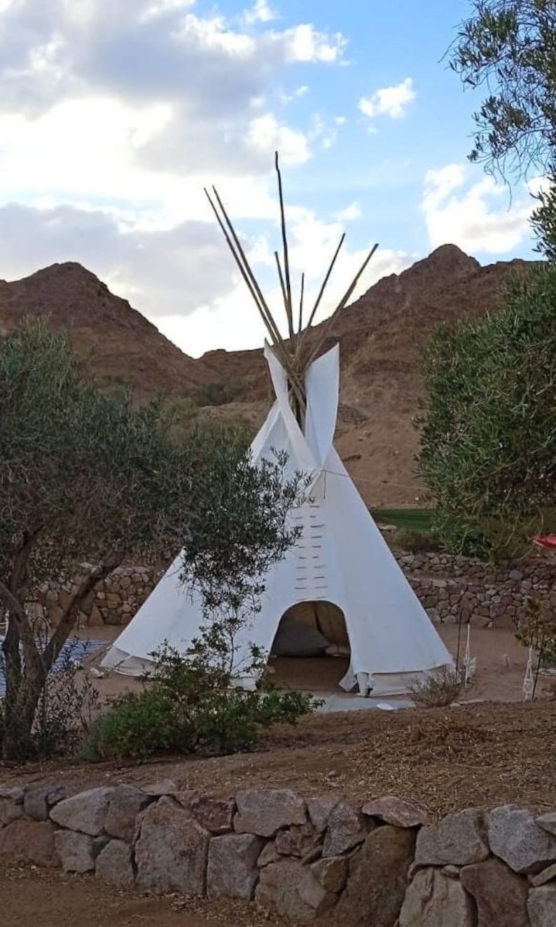 May include: A white teepee tent with a wooden frame, set up outdoors against a backdrop of brown hills and green trees.