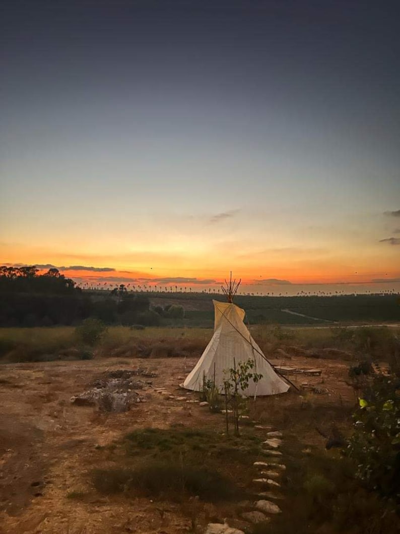 May include: A white teepee tent sits on a dirt path in a field with a sunset in the background.
