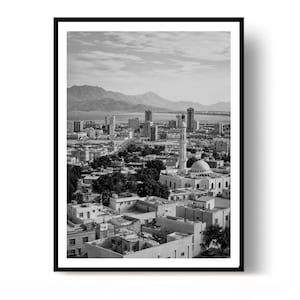 May include: A black and white framed print featuring an elevated cityscape view. The scene includes a prominent mosque with a tall minaret and dome in the foreground, surrounded by low-rise buildings, distant skyscrapers, and mountains under a cloudy sky.