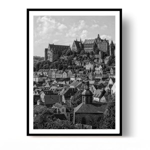 May include: A black and white photograph of a hillside town featuring a large castle complex at the peak. The scene displays numerous multi-story buildings with gabled roofs, dense trees, and a prominent church tower in the foreground under a cloudy sky.