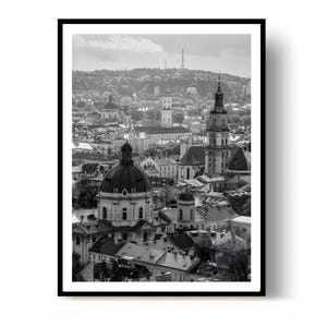May include: A black and white photographic print featuring an elevated cityscape view of Lviv, Ukraine. The scene highlights historic architecture, including a prominent domed church, multiple church spires, and dense urban rooftops.