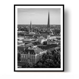 Op de afbeelding: Een zwart-wit ingelijste wandkunstprint met een verhoogd stadsgezicht. De scène toont dichte stedelijke architectuur, meerdere kerktorens die boven de daken uitsteken en omliggende bomen onder een heldere, lichte lucht.