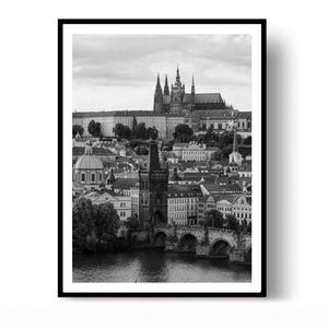 Op de afbeelding: Een zwart-wit fotoprint met een stadsgezicht van Praag. De afbeelding toont de rivier de Moldau op de voorgrond, de Karelsbrug met zijn stenen bogen en het historische complex van de Praagse Burcht gelegen op de heuvel boven de stad.