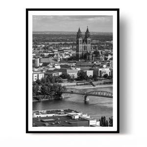 Może przedstawiać: Czarno-biały wydruk w ramie przedstawiający widok z lotu ptaka na miasto Magdeburg w Niemczech. Obraz ukazuje charakterystyczną katedrę z dwiema wieżami wznoszącą się nad panoramą miasta, most nad rzeką oraz okoliczne budynki.