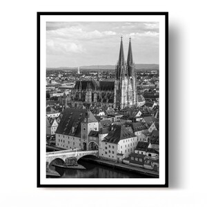 May include: A black and white photographic print featuring an elevated cityscape view of the Regensburg Cathedral with its twin spires, the Stone Bridge spanning the Danube River, and surrounding historic buildings under a cloudy sky.