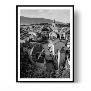 May include: A black and white photograph of a European town featuring a canal winding through stone buildings with pitched roofs. A prominent church spire rises in the background against a mountain range under a sky with light clouds.