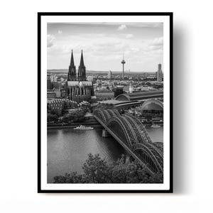 Könnte beinhalten: Eine Schwarz-Weiß-Fotografie des Kölner Doms mit seinen zwei Türmen, der Hohenzollernbrücke über den Rhein und der Skyline der Stadt im Hintergrund, alles eingerahmt von einem schlichten schwarzen Rahmen auf weißem Hintergrund.
