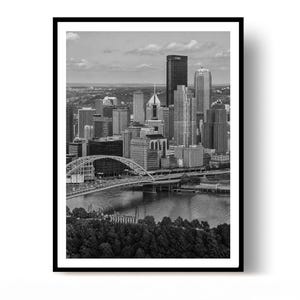 May include: A black and white photographic print of the Pittsburgh skyline featuring the Fort Pitt Bridge spanning the Monongahela River, surrounded by dense urban skyscrapers and a foreground of trees, all framed within a simple black border.