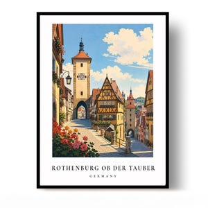 May include: A framed art print featuring a sunny street scene in Rothenburg ob der Tauber, Germany. The image displays half-timbered buildings, a stone clock tower, and vibrant red and pink flowers in the foreground under a blue sky.