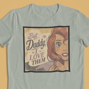 May include: A light green t-shirt featuring a graphic of a cartoon woman with red hair and the text "But Daddy, I Love Them." The graphic is in a black rectangle with a vintage style.