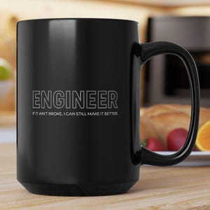 Puede incluir: Una taza de cerámica negra con un asa grande. La taza presenta la palabra "ENGINEER" en letras mayúsculas blancas, con la frase "IF IT AIN'T BROKE, I CAN STILL MAKE IT BETTER" debajo. La taza está sobre una superficie de madera.