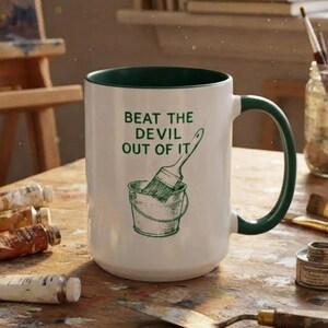 May include: A white ceramic mug with a dark green interior and handle. The front features a dark green graphic of a paint bucket and brush with the text "BEAT THE DEVIL OUT OF IT" printed in dark green capital letters above the illustration.