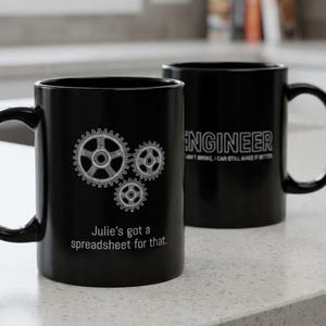 Może przedstawiać: Dwa czarne ceramiczne kubki z białym tekstem i grafikami. Jeden kubek posiada trzy srebrne ilustracje kół zębatych oraz napis "Julie's got a spreadsheet for that." Drugi kubek zawiera tekst "ENGINEER" oraz "AIN'T BROKE, I CAN STILL MAKE IT BETTER."