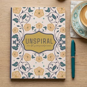 May include: A journal with a floral design and the word "UNSPIRAL" in a gold frame. A teal pen and a cup of coffee sit beside it. The journal's cover has a cream background with beige flowers and teal leaves.