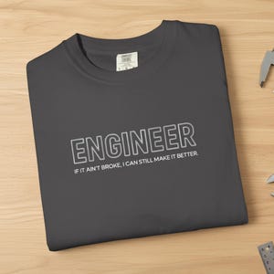 May include: A dark gray crewneck t-shirt featuring the white text "ENGINEER" and "IF IT AIN'T BROKE, I CAN STILL MAKE IT BETTER." The shirt is displayed alongside a metal caliper, a gear, pliers, an adjustable wrench, a ruler, and a notebook.