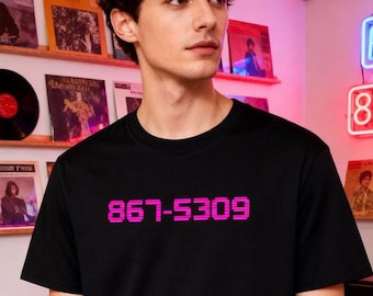 May include: A black t-shirt with the magenta text "867-5309" is worn by a person in a record store. The store has shelves of vinyl records and neon signs. The person is wearing black pants.