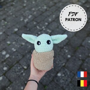 Peut inclure: Un jouet en peluche vert et marron crocheté ressemblant à un personnage de la franchise Star Wars. Le jouet est assis dans un pot crocheté marron. L'image comprend le texte "PDF PATRON".