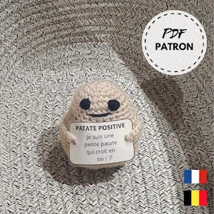 Puede incluir: Patrón de crochet amigurumi para un personaje de patata que sostiene un cartel que dice "Patate Positive" y "Je suis une petite patate qui croit en toi!"  El cartel es blanco con texto negro y un corazón. La patata es beige con ojos negros y una sonrisa. El patrón está disponible para descargar en PDF.