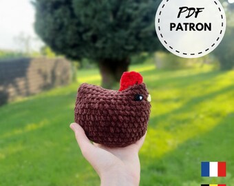 May include: A brown crocheted chicken plush toy with a red comb and yellow beak. The toy is being held by a hand in front of a green grassy background. The text "PDF PATRON" is visible in a white circle with a black dashed border.