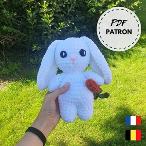 May include: White crocheted bunny rabbit plush toy holding a carrot. The bunny has black eyes and a pink nose. The toy is being held by a hand wearing a black bracelet. The image includes the text "PDF PATRON".
