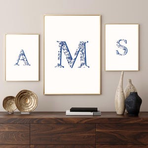 Puede incluir: Tres impresiones enmarcadas con las letras "A", "M" y "S" en patrones florales azules sobre un fondo blanco. Los marcos tienen bordes dorados. Las impresiones se exhiben sobre un mueble de madera oscura.