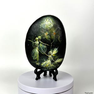 Puede incluir: Una piedra pintada a mano con tres hadas bailando en un bosque verde oscuro con flores blancas. La piedra está sobre un soporte negro y una superficie blanca.