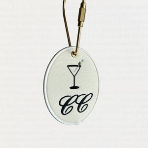 Könnte beinhalten: Ein ovales, cremefarbenes, besticktes Etikett mit einem Martini-Glas-Design und den Buchstaben "CC" in schwarzer Schrift. Das Etikett hat einen goldfarbenen Metallverschluss und einen weißen Rand. Ein kleiner Zweig aus grüner und weißer Stickerei akzentuiert das Martini-Glas.