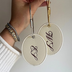 Könnte beinhalten: Zwei cremefarbene ovale Gepäckanhänger mit gestickten Initialen in dunkler brauner Schrift. Jeder Anhänger hat einen silber- oder goldfarbenen Metallverschluss. Die Anhänger werden von einer Person gehalten.