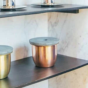 Könnte beinhalten: Zwei Metallbehälter mit Deckeln auf einem schwarzen Regal. Der Behälter links ist golden und der Behälter rechts ist kupferfarben.