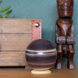 May include: A striped agate sphere on a wooden stand. The sphere is a deep purple with white stripes. The sphere is sitting on a wooden table in front of a wooden tiki statue.
