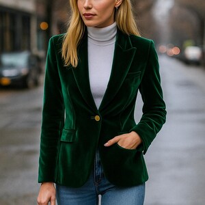 May include: A dark green velvet blazer with a single gold button closure. The blazer has a classic tailored design with a notched lapel and front pockets. The person is wearing a light gray turtleneck and blue jeans.
